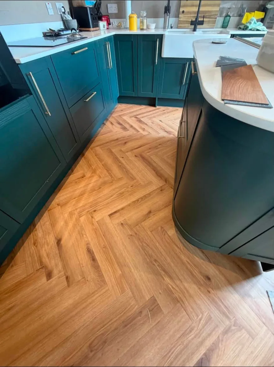 Warm herringbone wood-effect floor fitted in a kitchen with dark green shaker units and a curved island
